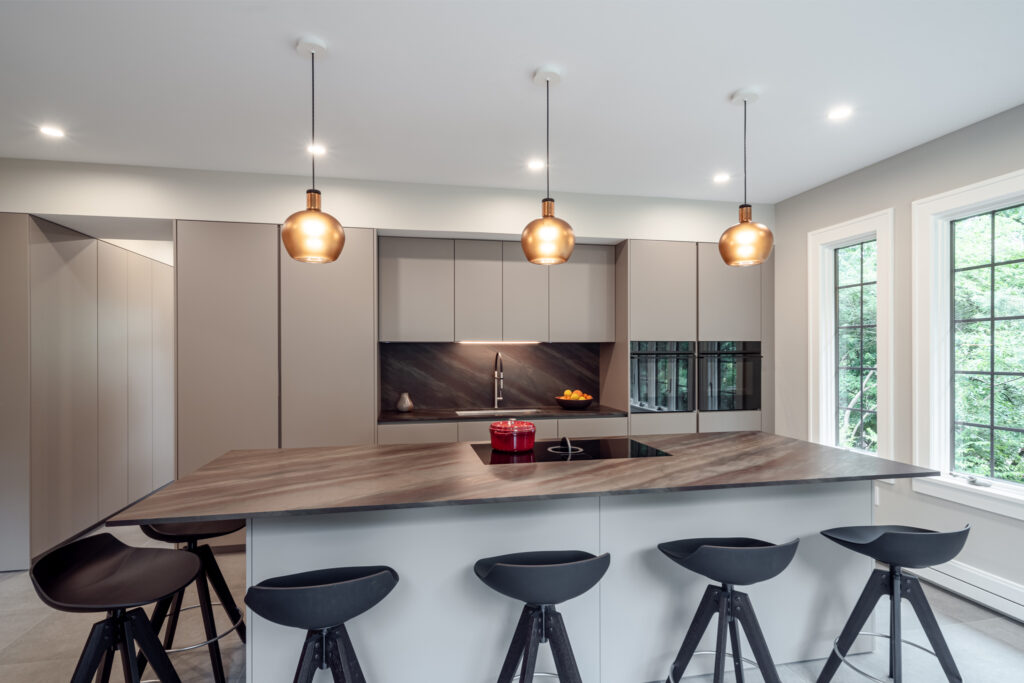 An award-winning kitchen featuring a sleek design with integrated lighting. The space includes a modern island with a wood countertop, surrounded by stylish black stools. Three pendant lights hang above the island, enhancing the contemporary aesthetic while providing functional illumination.