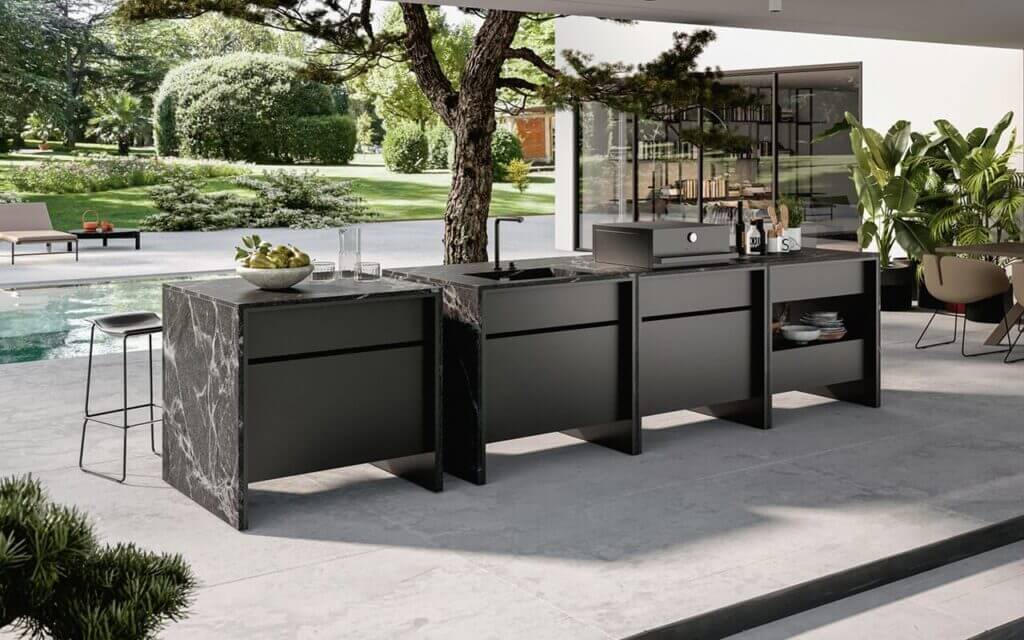 An example of NOLI's luxury outdoor kitchens, featuring a freestanding stainless steel modular kitchen island clad in black stone with wood accents, showcasing weather-resistant materials in a lush poolside garden.