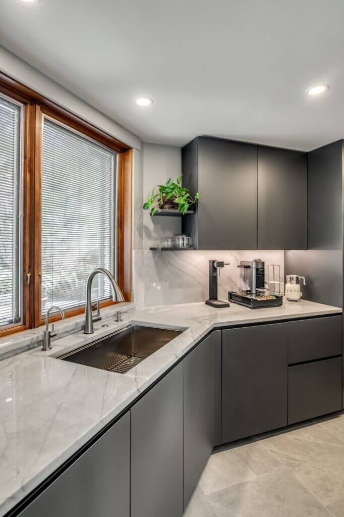 Corner view of a modern kitchen with dark matte cabinetry, white marble countertops, an undermount stainless steel sink, and natural light streaming through large windows with wooden trim. A small potted plant adds a touch of greenery above the counter.