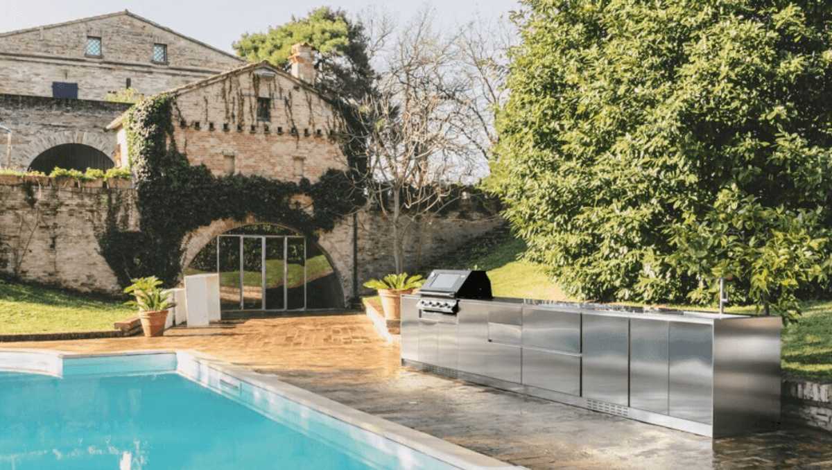 Stainless steel Italian outdoor kitchen beside a pool with stone architecture in the background.